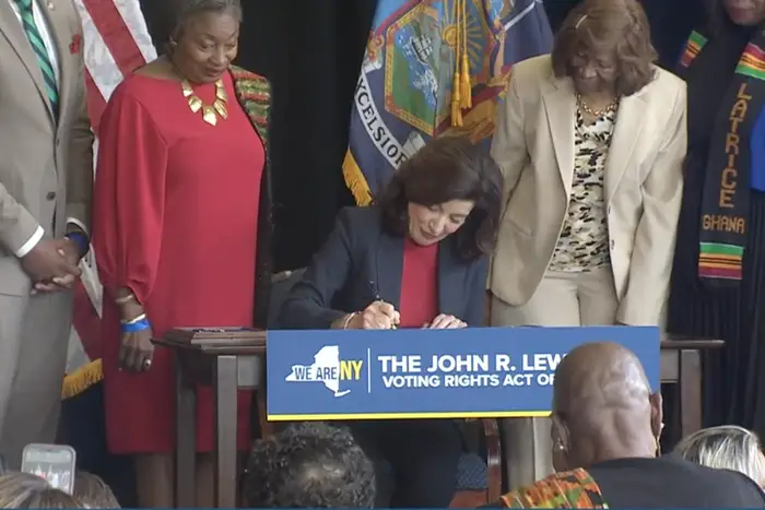 Gov. Kathy Hochul is seated and surrounded by lawmakers and activists as she signs the John R. Lewis Voting Rights Act of New York into law.